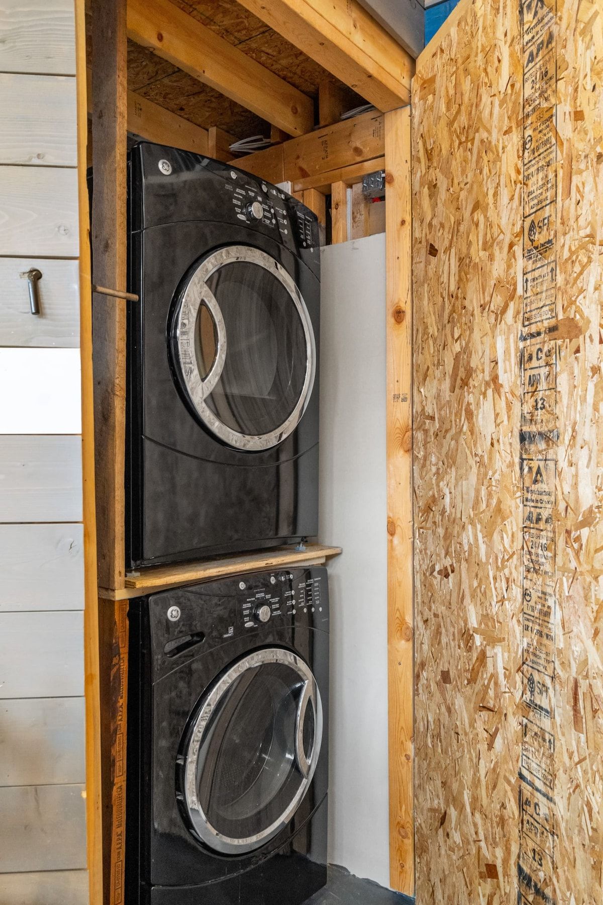 A compact laundry room with two stacked washing machines, wooden walls, and a visible door handle.