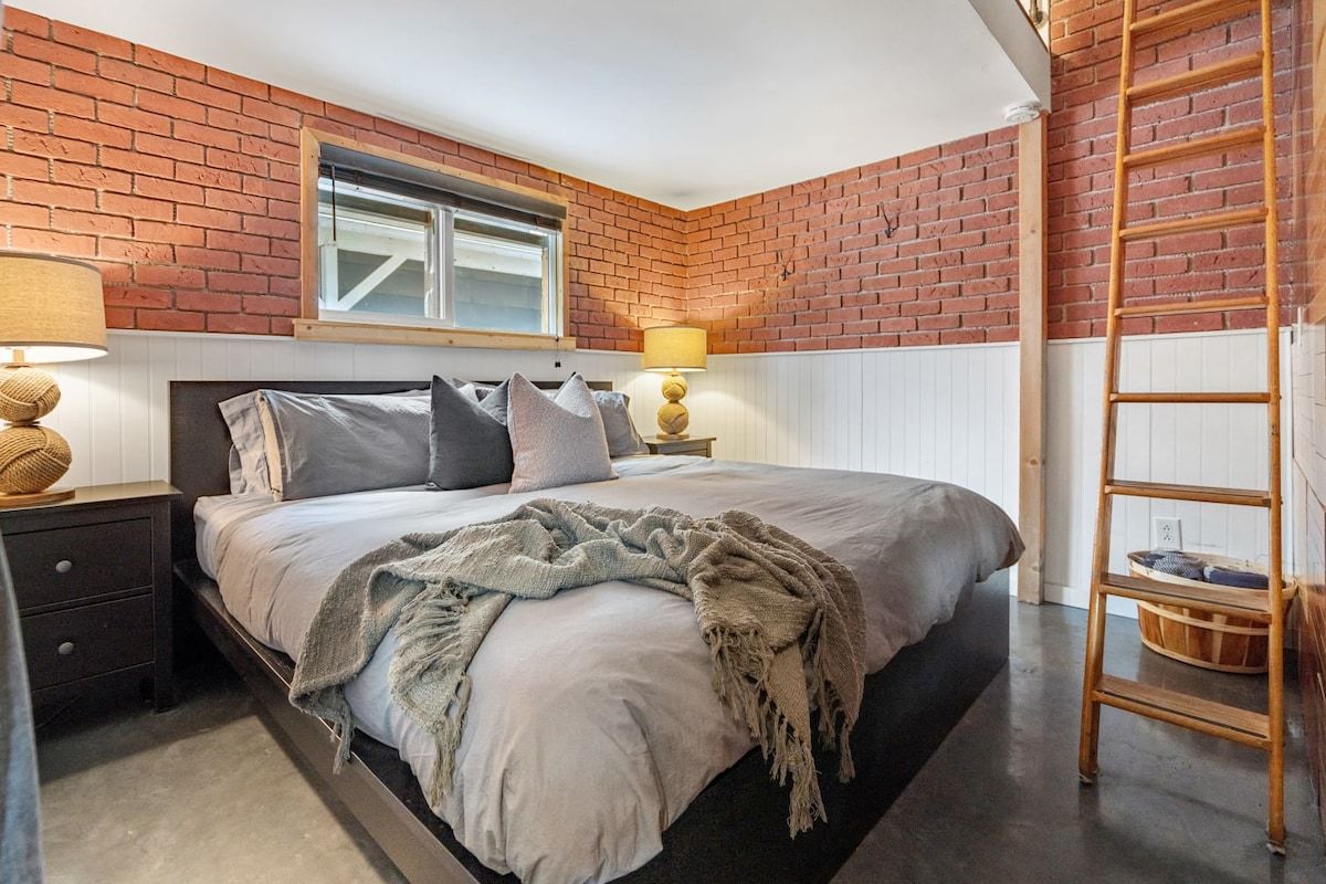 Une chambre avec un mur en briques, un lit avec des oreillers, une table de chevet, une lampe et une échelle.