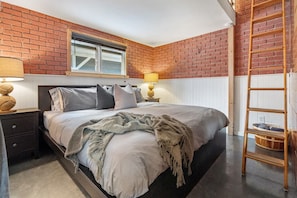 A bedroom with a brick wall, a bed with pillows, a nightstand, a lamp, and a ladder. - Historic Firehall Guesthouse with Private Covered Patio & Wood Stove (Rosebud)