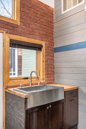 A kitchen with a wooden window frame, a stainless steel sink, and dark wooden cabinets. - Historic Firehall Guesthouse with Private Covered Patio & Wood Stove (Rosebud)