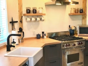 A kitchen with a sink, stove, oven, and shelves with jars and mugs.