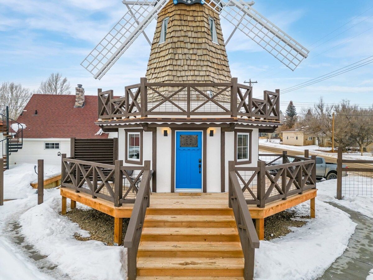Eine Windmühle mit einer blauen Tür, Holztreppen und einem Metallzaun.