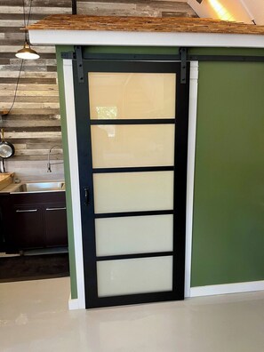 A sliding door with a frosted glass panel and a black frame, leading to a kitchen area with a wooden backsplash and a hanging pendant light.