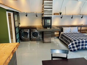 A compact room with a bed, a washing machine, a dryer, and a small table.