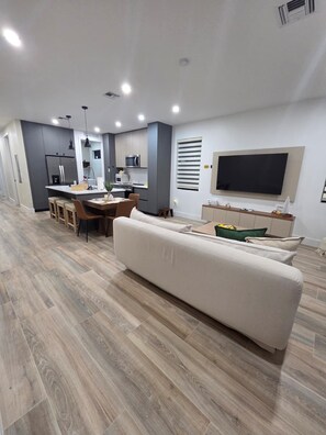 A modern living room with a large white sofa, a dining area with a wooden table and chairs, a kitchen with grey cabinets and stainless steel appliances, and a flat-screen TV mounted on the wall. - Marvelous 4-bedroom House Near Beaches (Hollywood)