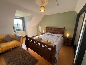 Ein Schlafzimmer mit einem hölzernen Bett, einem kleinen Nachttisch mit Lampe, einem Fenster mit Vorhängen und einem gelben Sofa.