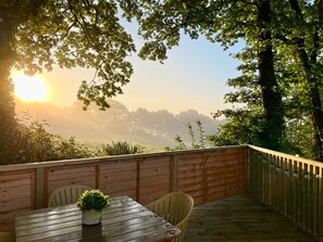 Eine Holzterrasse mit Tisch und Stühlen, mit Blick auf eine neblige Landschaft mit Bäumen und einem Sonnenaufgang.