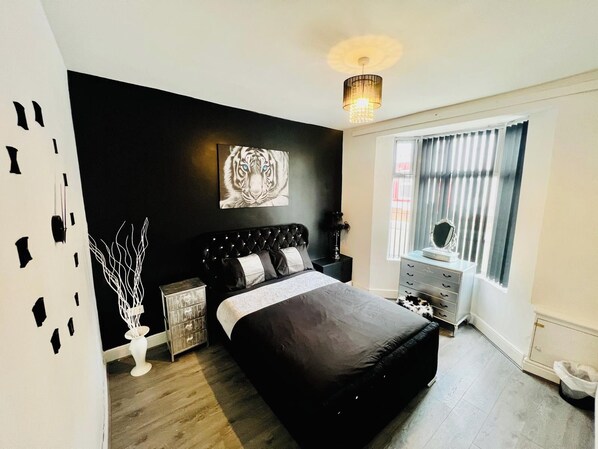 A bedroom with a black bed, a white wall with a tiger print, a window with blinds, and a bedside table with a mirror.
