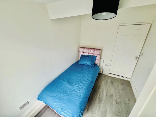 Una habitación pequeña con cama individual, cabecera rosa, colcha azul y puerta blanca.