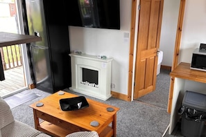 A room with a fireplace, a television mounted on the wall, a wooden table with a black object on it, and a door leading to another room. - Cabin Sleeps 2 with Heating (Mablethorpe)