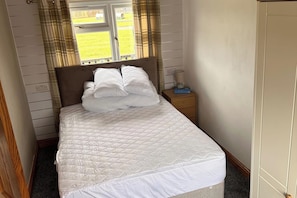 A neatly made bed with a quilted white duvet, a wooden nightstand with a lamp, and a window with curtains. - Cabin Sleeps 2 with Heating (Mablethorpe)