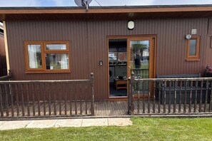 A wooden building with a glass door entrance, a small porch with a railing, and a satellite dish on the roof. - Cabin Sleeps 2 with Heating (Mablethorpe)