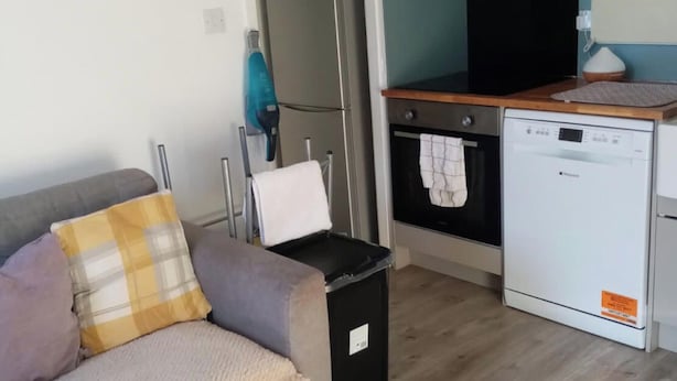 Een compacte woonruimte met een kitchenette, een bank met kussens, een prullenbak en een wasmachine.