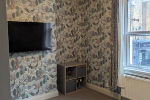 A room with a patterned wallpaper, a flat-screen TV mounted on the wall, a small wooden cabinet, and a window with a view of buildings outside. - Central Stratford Gem (London)