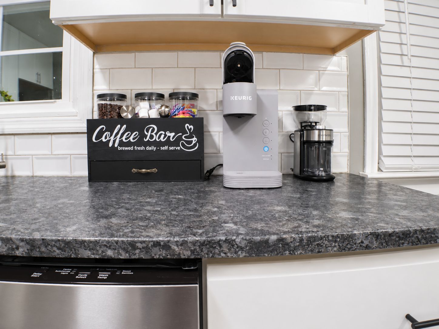 Disposición de una barra de café con una máquina Keurig, un molinillo de café y una cafetera sobre una encimera de granito.