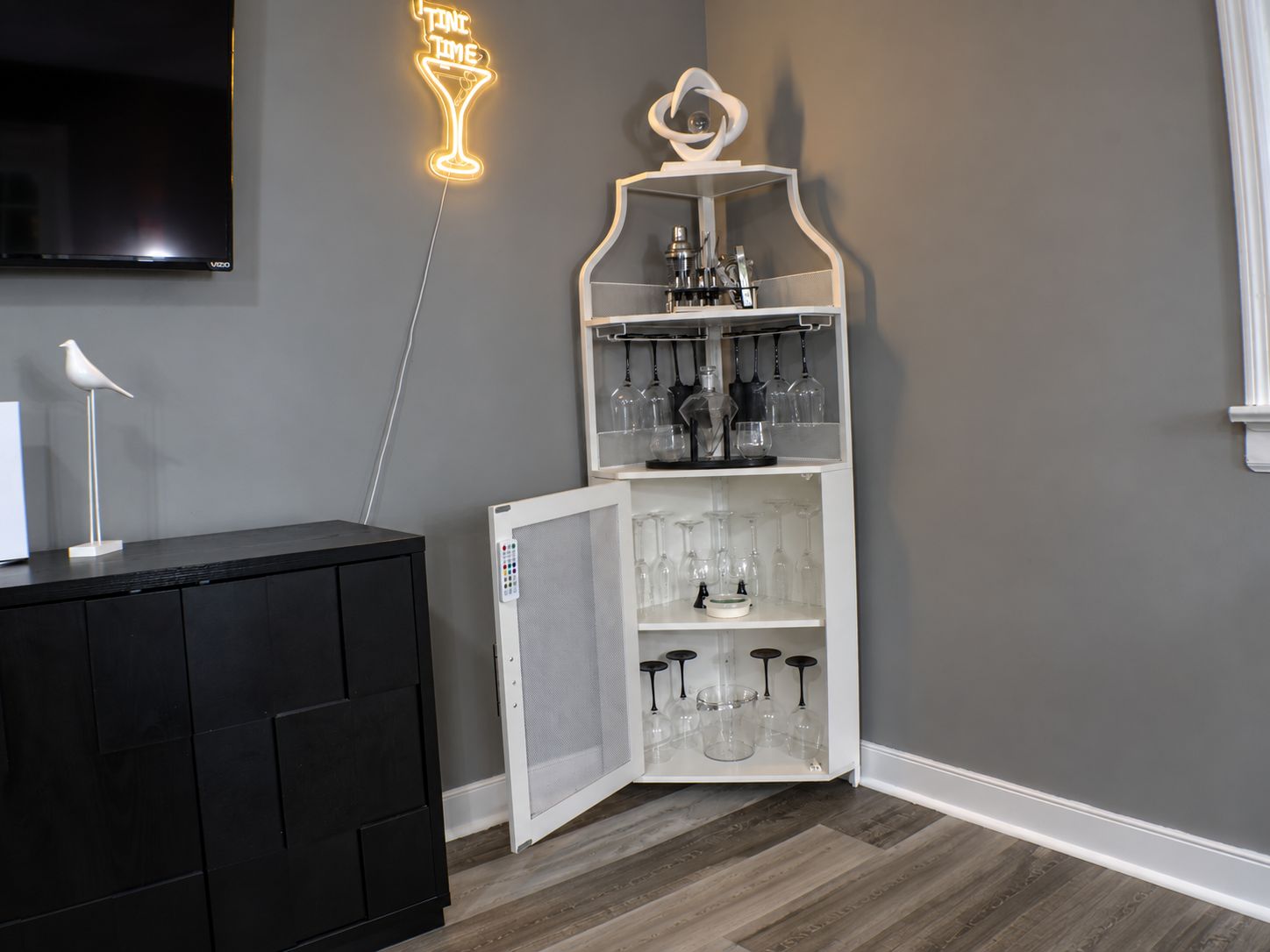 Een kamer met een wandgemonteerde flatscreen-tv, een neonbord, een wit kastje met wijnglazen en een zwart kastje met een witte lamp.