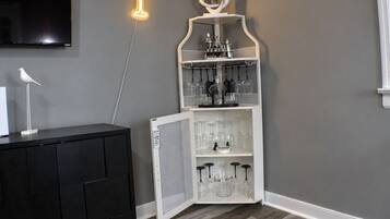 Een kamer met een wandgemonteerde flatscreen-tv, een neonbord, een wit kastje met wijnglazen en een zwart kastje met een witte lamp.