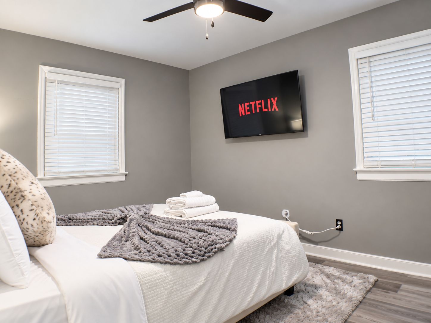 A bedroom with a bed, a ceiling fan, a wall-mounted TV displaying 'NETFLIX', and a window with blinds.
