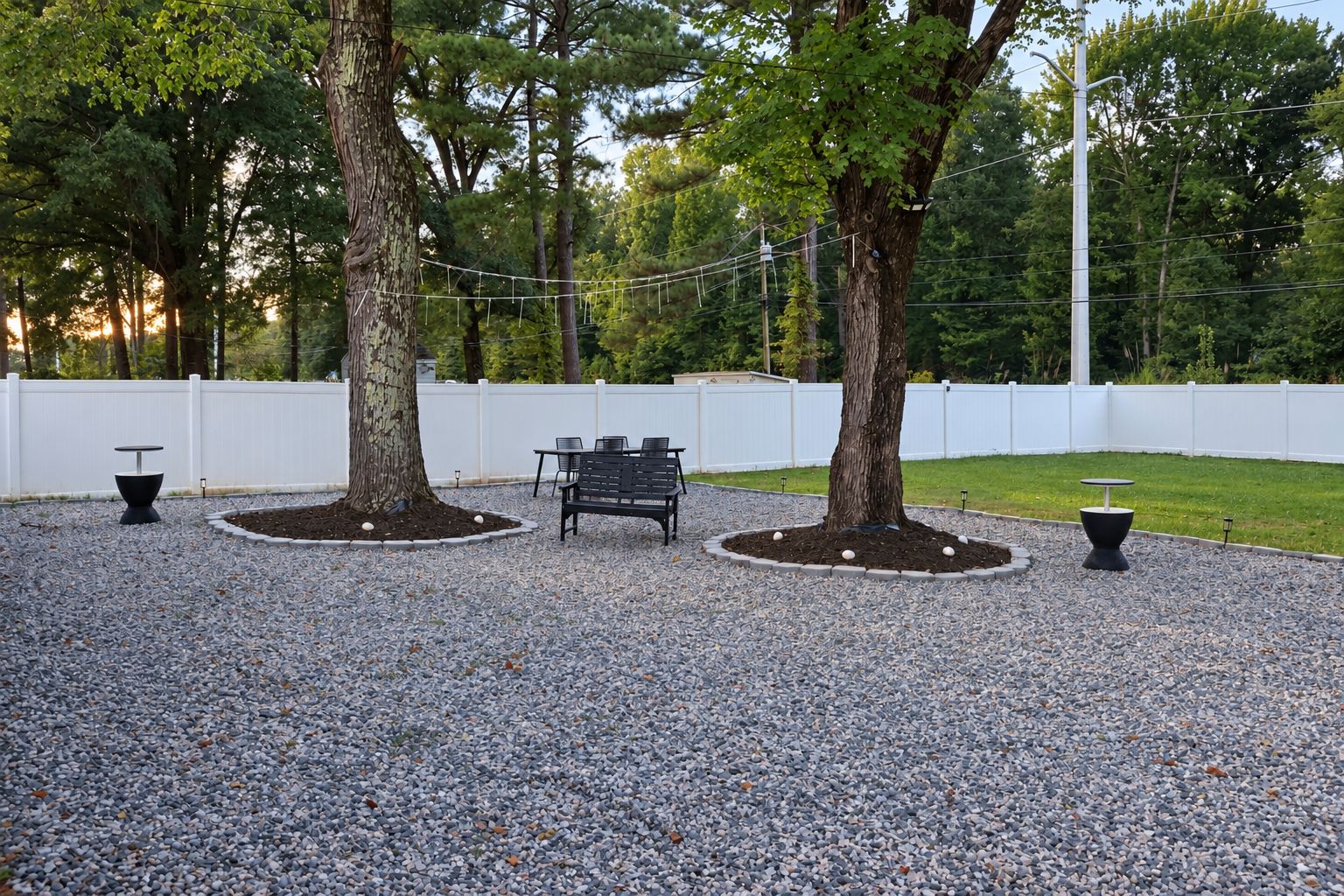 A gravel patio with a bench and two tables, surrounded by trees and a white fence.