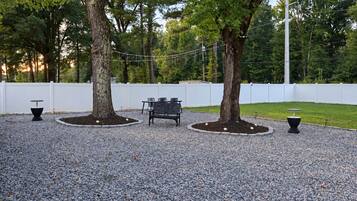 A gravel patio with a bench and two tables, surrounded by trees and a white fence.