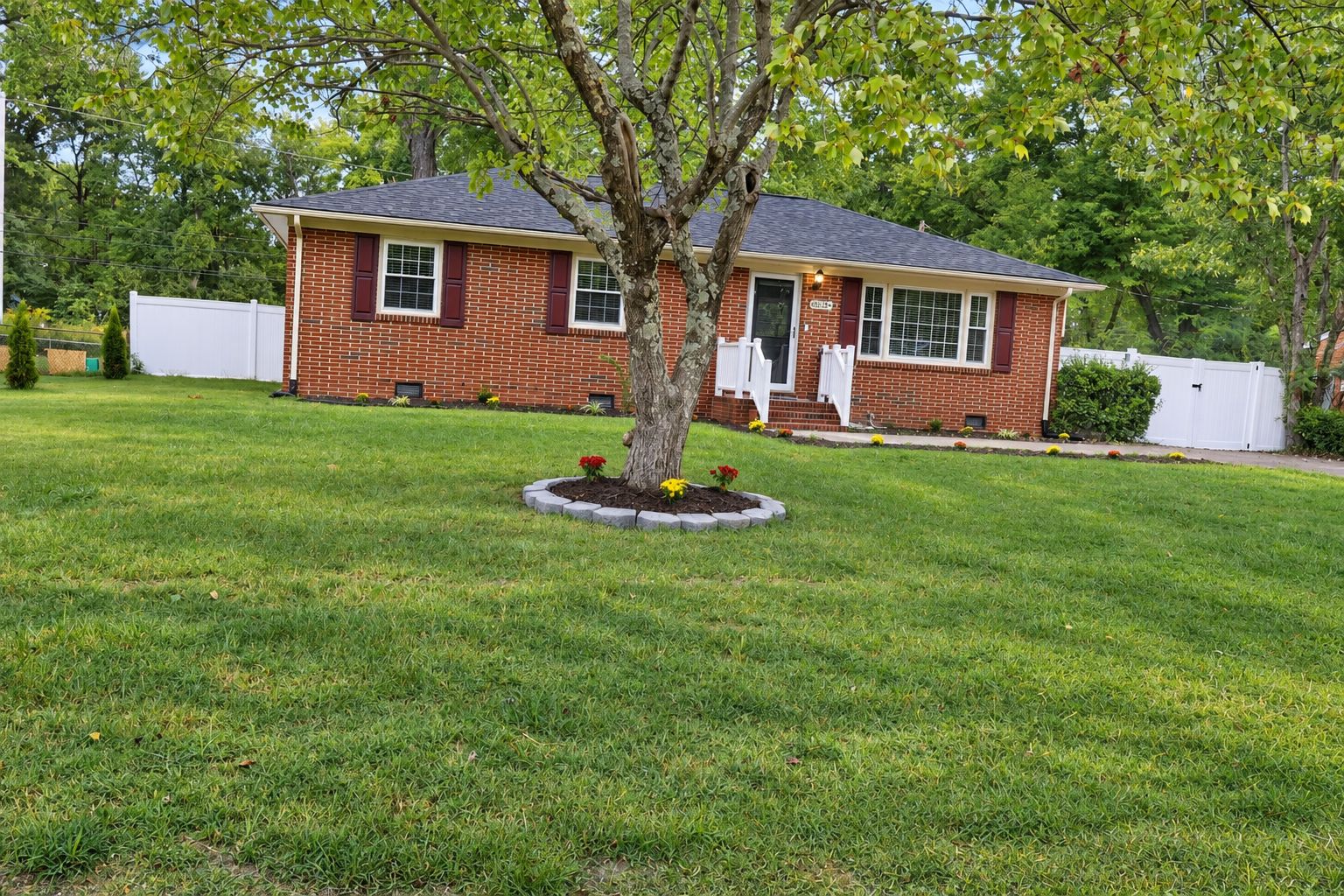 A single-story brick house with a front porch, surrounded by a well-maintained lawn and a tree.
