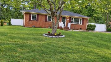 A single-story brick house with a front porch, surrounded by a well-maintained lawn and a tree.