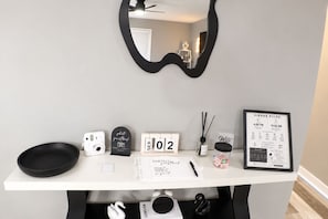 A hotel reception desk with a black-framed mirror, a framed 'Airbnb Rules' sign, a calendar, a photo booth sign, a small black bowl, a white camera, a white pen, a small bottle, and a black swan decoration.