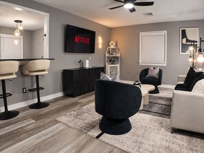 Una sala de estar moderna con un sofá negro, un sillón blanco, una mesa de centro negra y un mueble negro para televisor con el logo de Netflix.