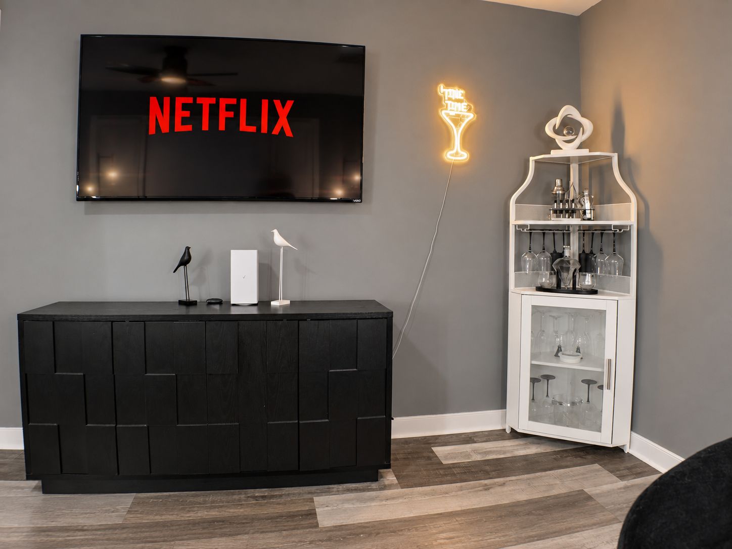 Un salon moderne avec un meuble de rangement noir pour l’équipement audiovisuel, un téléviseur fixé au mur affichant le logo « NETFLIX », un néon et un meuble blanc contenant de la verrerie.