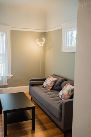 Ein Wohnzimmer mit einem grauen Sofa, zwei Zierkissen, einem schwarzen Couchtisch und einer Stehlampe mit einer herzförmigen Leuchte.