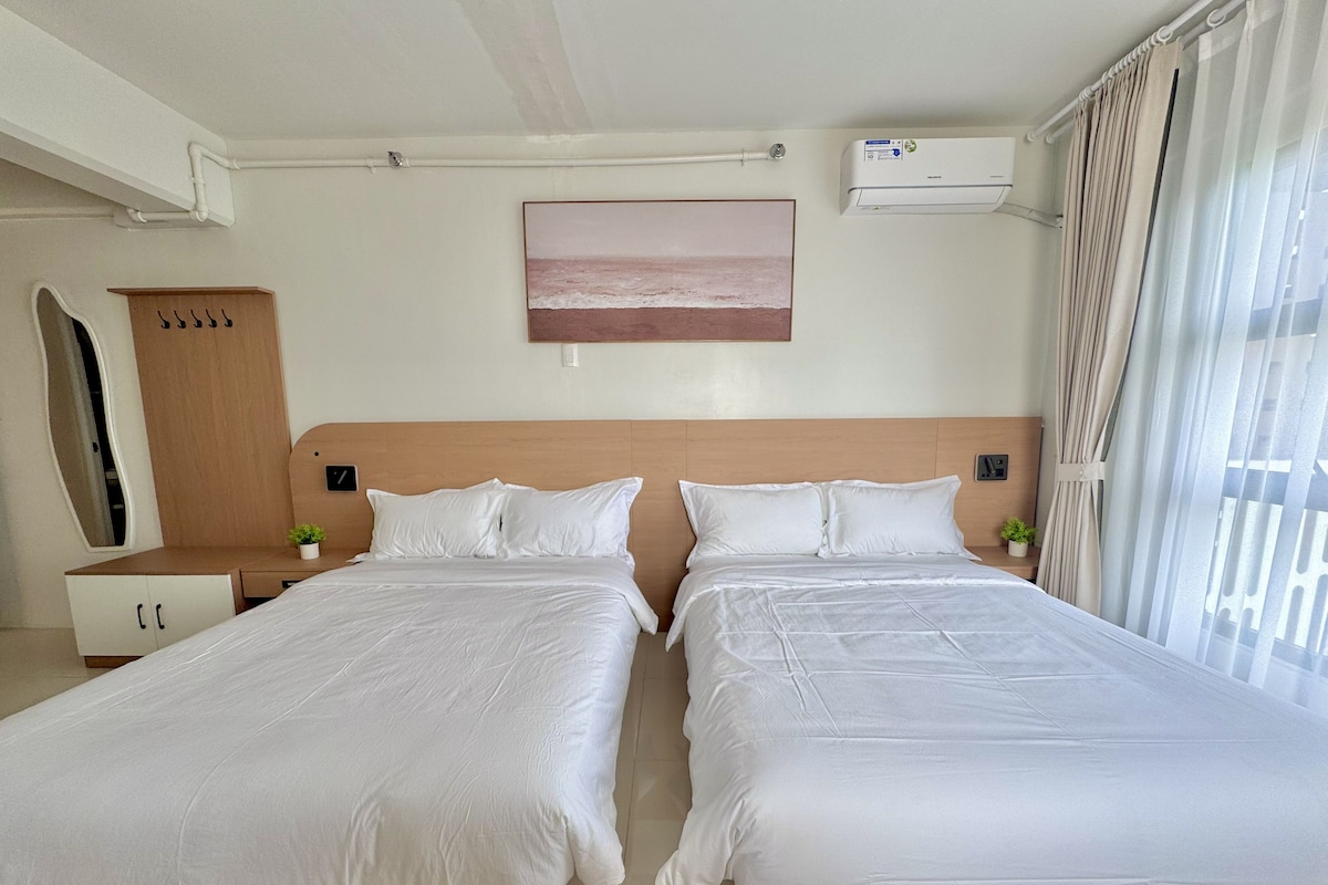 A hotel room with two beds, a wooden headboard, a small bedside table with a plant, a wall-mounted air conditioner, and a window with curtains.