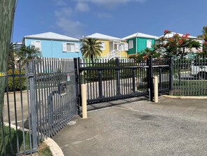 A gated entrance with a metal fence, a palm tree, and colorful buildings in the background. - Relaxing Gated Condo w/ Huge Pool, Walk to Beach (Christiansted)