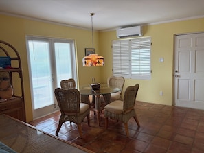 A dining area with a glass table surrounded by wicker chairs, a hanging light fixture, and a door with a window. - Relaxing Gated Condo w/ Huge Pool, Walk to Beach (Christiansted)