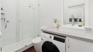 Un baño moderno con ducha envuelta en vidrio, un lavamanos blanco y una lavadora empotrada.