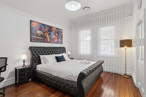 A bedroom with a tufted headboard bed, a bedside table with a lamp, a chair, a large abstract painting, sheer curtains, and a floor lamp.