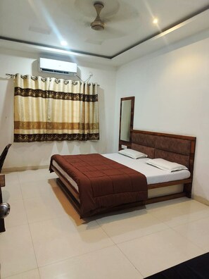 A bedroom with a bed, a ceiling fan, an air conditioning unit, a window with curtains, and a mirror. - HOTEL MANSAROVAR (Omkareshwar)