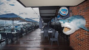 Área de refeições ao ar livre com mesas e cadeiras, vista de barcos ao fundo e uma decoração de tubarão na parede.