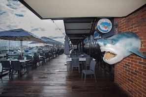 Un área de comedor al aire libre con mesas y sillas, vista de barcos al fondo y una decoración de tiburón en la pared.