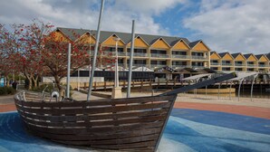 Hotel à beira-mar com várias varandas, uma escultura em forma de barco em primeiro plano e uma ponte para pedestres sobre a água.