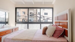 Quarto com cama, janela, ventilador de teto e vista para prédios.