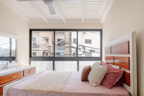 Una camera da letto con un letto, una finestra, una ventola a soffitto e vista sugli edifici circostanti.