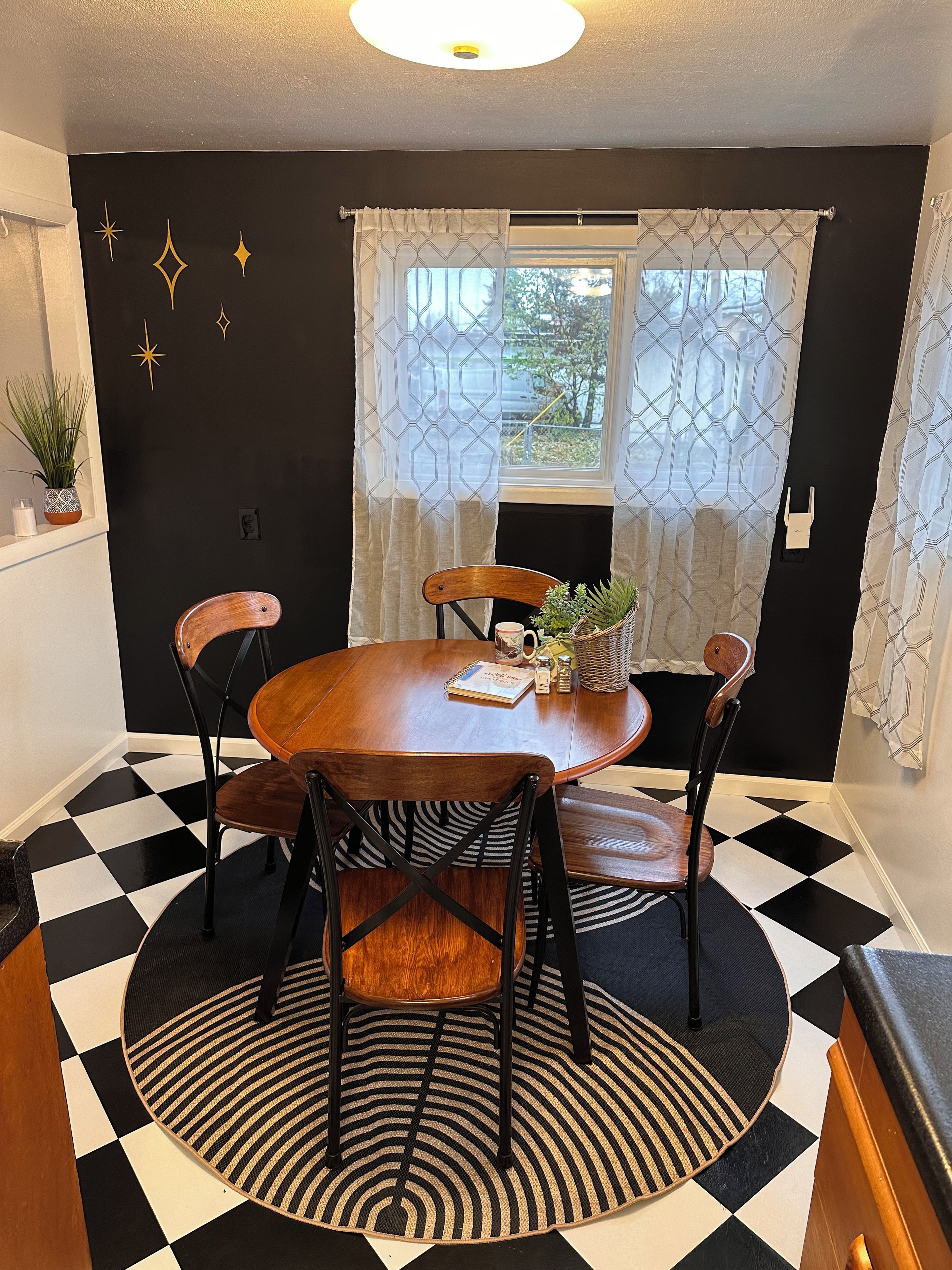 A dining area with a round table, four chairs, a patterned rug, and a window with patterned curtains.