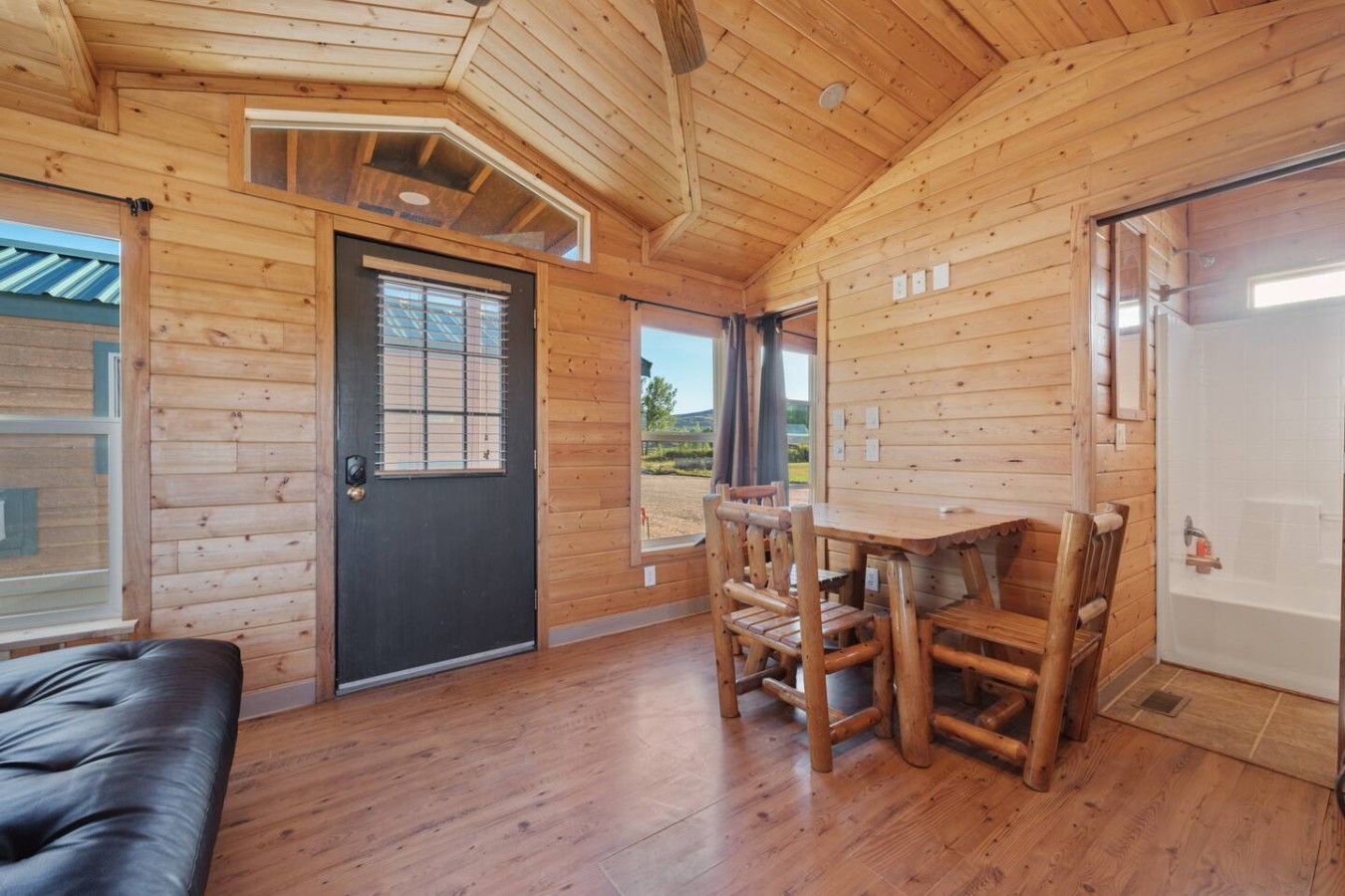 L’intérieur d’un chalet en bois, avec une porte noire, une table à manger entourée de chaises et une vue sur l’extérieur, visible à travers une fenêtre.