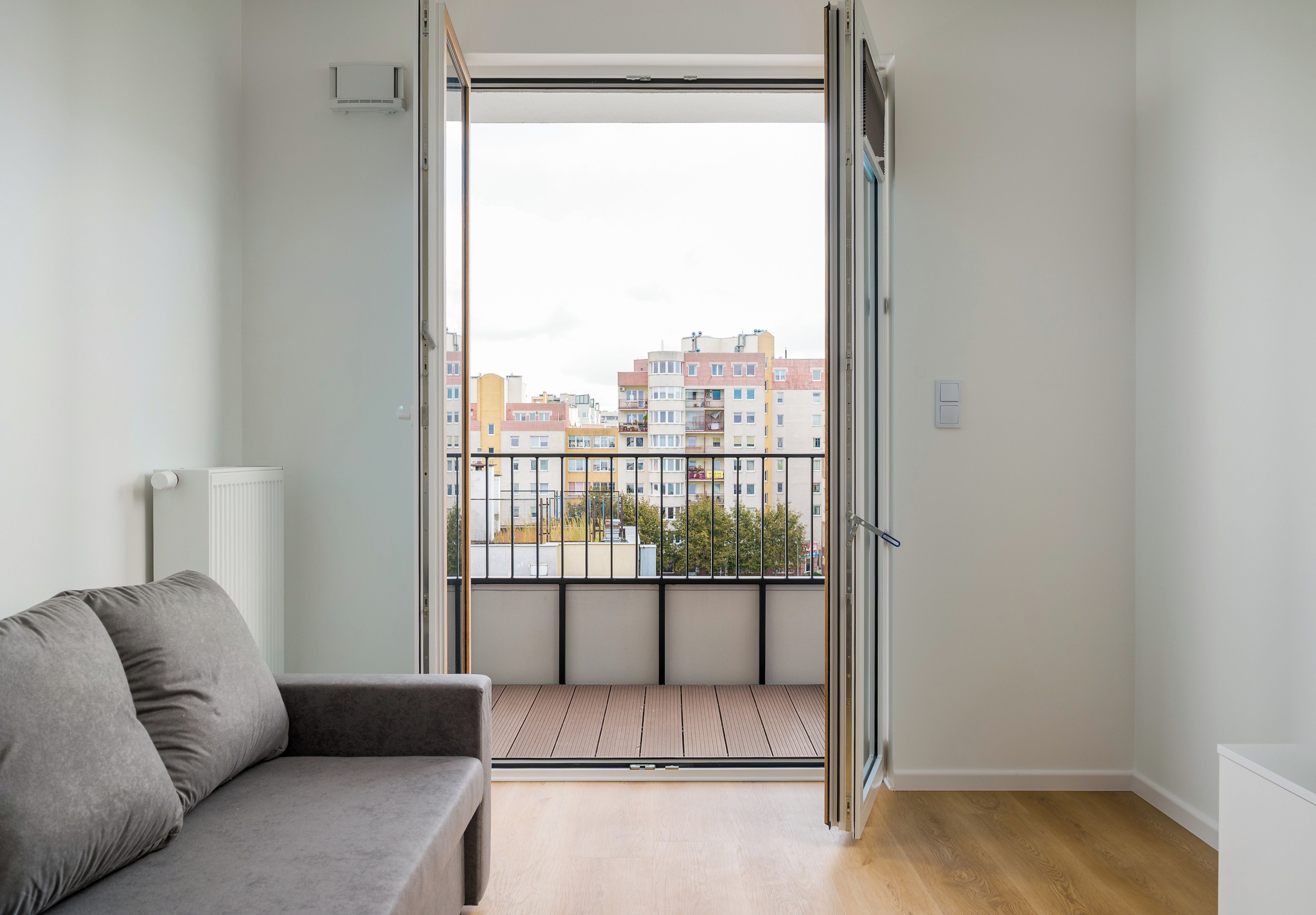 Une pièce avec un canapé gris, un radiateur, une fenêtre donnant sur des immeubles et une porte.