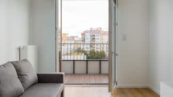 Une pièce avec un canapé gris, un radiateur, une fenêtre donnant sur des immeubles et une porte.