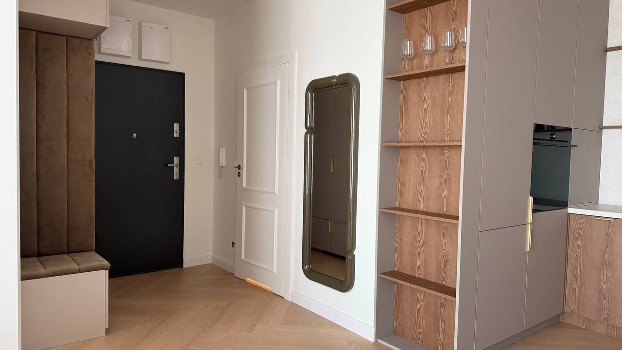 Un appartement moderne avec un banc en bois, une porte sombre, un placard à miroir et une cuisine dotée d’étagères en bois.