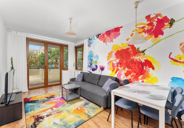 Ein modernes Wohnzimmer mit einem grauen Sofa, einem Esstisch mit Stühlen, einem farbenfrohen abstrakten Gemälde an der Wand und einem großen Fenster mit Blick auf Grünflächen.