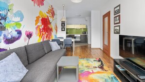 Una sala moderna con un sofá gris, una mesa de centro, un cuadro abstracto colorido, un televisor de pantalla plana y una cocina al fondo.