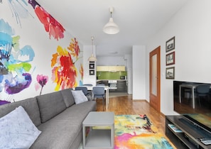 Ein modernes Wohnzimmer mit grauem Sofa, einem Couchtisch, einem farbenfrohen abstrakten Gemälde, einem Flachbildfernseher und einer Küche im Hintergrund.