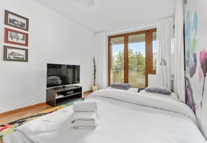 Ein modernes Hotelzimmer mit einem großen Bett, einem Flachbildfernseher, einem kleinen Tisch und einem Fenster mit Blick auf Bäume.